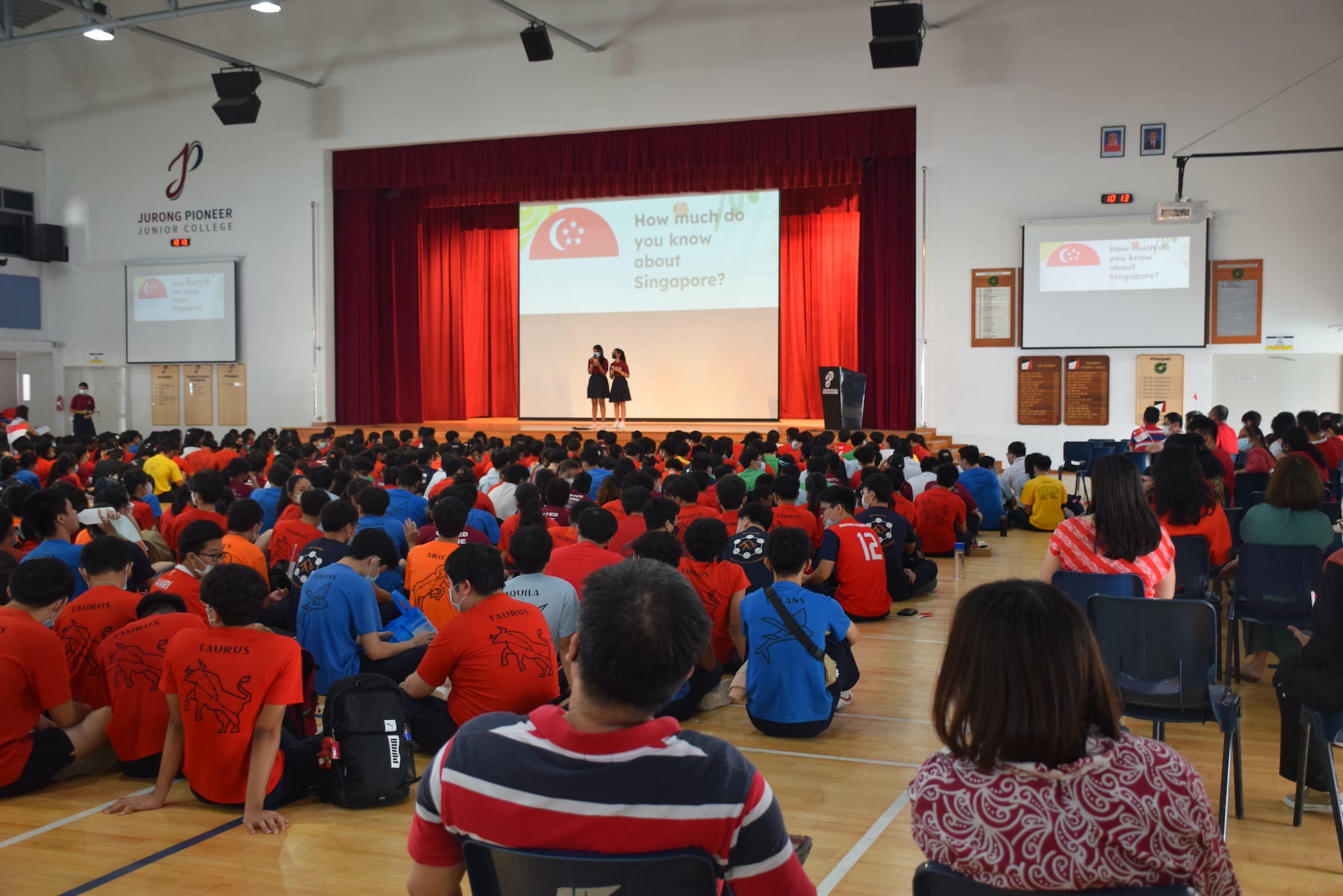 Students sit on a hall floor at Jurong Pioneer Junior College watching a Singapore quiz presentation on stage.